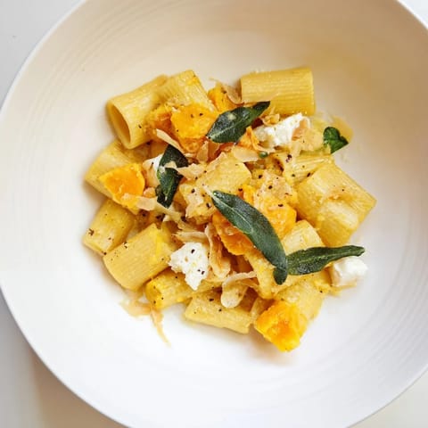 A vibrant close-up of creamy roasted butternut squash and sage goat cheese pasta, garnished with fresh herbs.