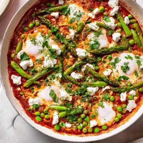 In a rustic skillet, Pea and Broad Bean Shakshuka features tender peas, broad beans, and asparagus simmered in a fragrant herb tomato sauce.
